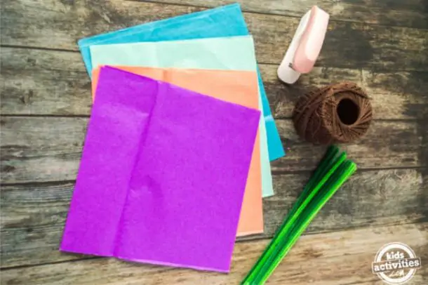 supplies for making pom pom mexican paper flowers like tissue paper, twine, stapler and pipe cleaners laid out on a wood surface