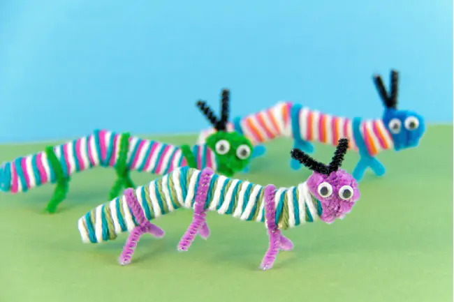 Caterpillar popsicle stick craft with standing caterpillars covered in yarn, pom pom heads, and pipe cleaner legs.